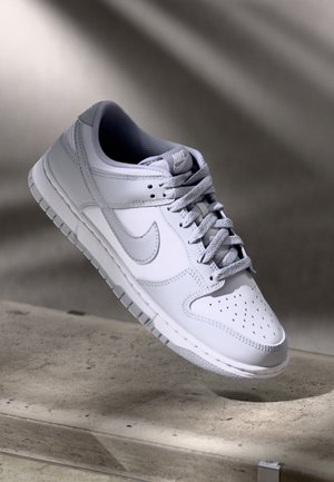 Biały sneaker Nike z szarymi sznurowadłami i logo Swoosh, przedstawiony unoszący się nad teksturowaną powierzchnią betonową, przy delikatnym oświetleniu i neutralnym tle.