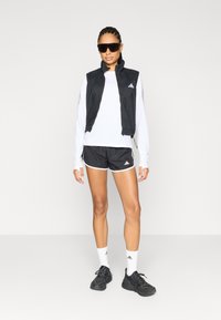 Chaleco negro sin mangas sobre una camisa blanca de manga larga, combinado con pantalones cortos negros con detalles en blanco y zapatos deportivos negros. Gafas de sol incluidas.