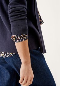 Henkilö, joka on pukeutunut tummansiniseen neuleeseen kukkakuosisen pitkähihaisen paidan päälle, hihojen ollessa käärittynä, yhdistettynä sinisiin farkkuihin.