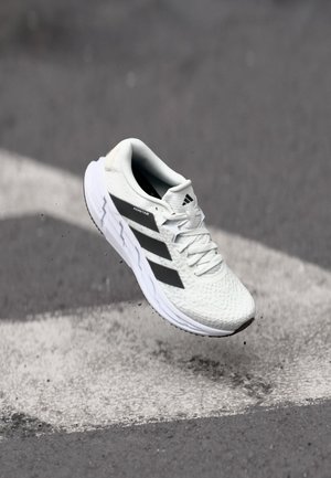 Biały but do biegania Adidas z czarnymi paskami w powietrzu nad asfaltem z białymi oznaczeniami drogowymi.
