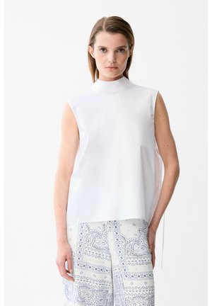 Femme aux cheveux bruns raides portant un haut blanc sans manches à col montant et un pantalon blanc à jambes larges avec un motif paisley bleu, debout sur fond blanc.