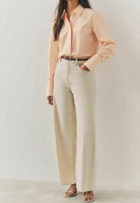 Chemise à manches longues rayée de couleur orange clair, associée à un pantalon beige taille haute à jambes larges, complété par une ceinture assortie et des sandales à bouts ouverts.