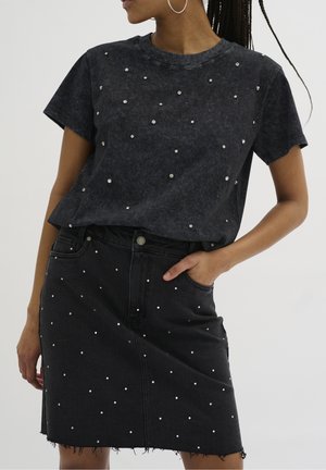 Kvinde iført en sort top med korte ærmer og matchende nederdel, begge pyntet med små sølvnitter, poserer med den ene hånd i lommen.