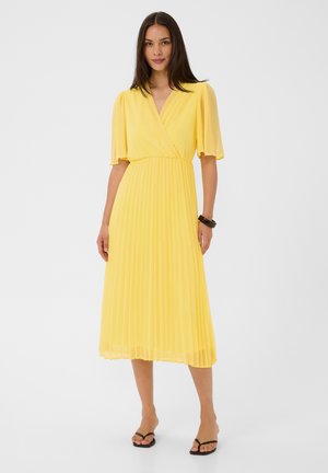 Femme portant une robe midi jaune vif avec une jupe plissée, des manches courtes à volants et des sandales noires sur un fond clair uni.