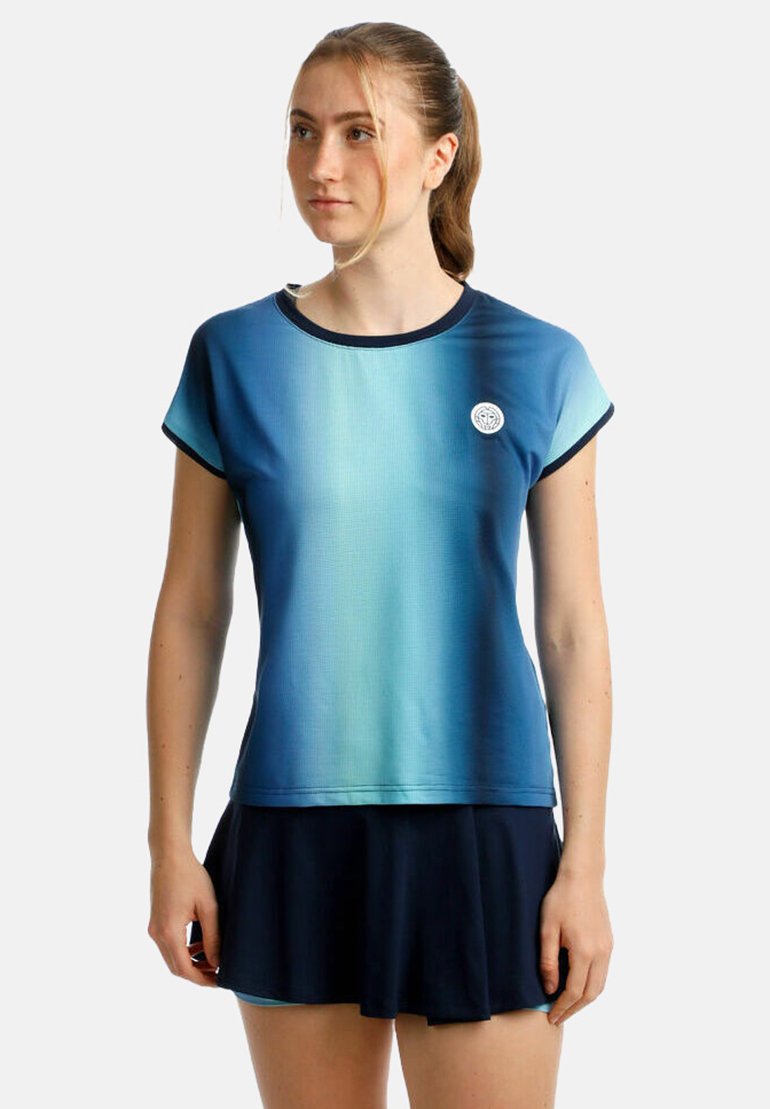 Joven mujer con una camiseta de manga corta de degradado azul y una falda negra con pliegues, de pie contra un fondo claro y sencillo.