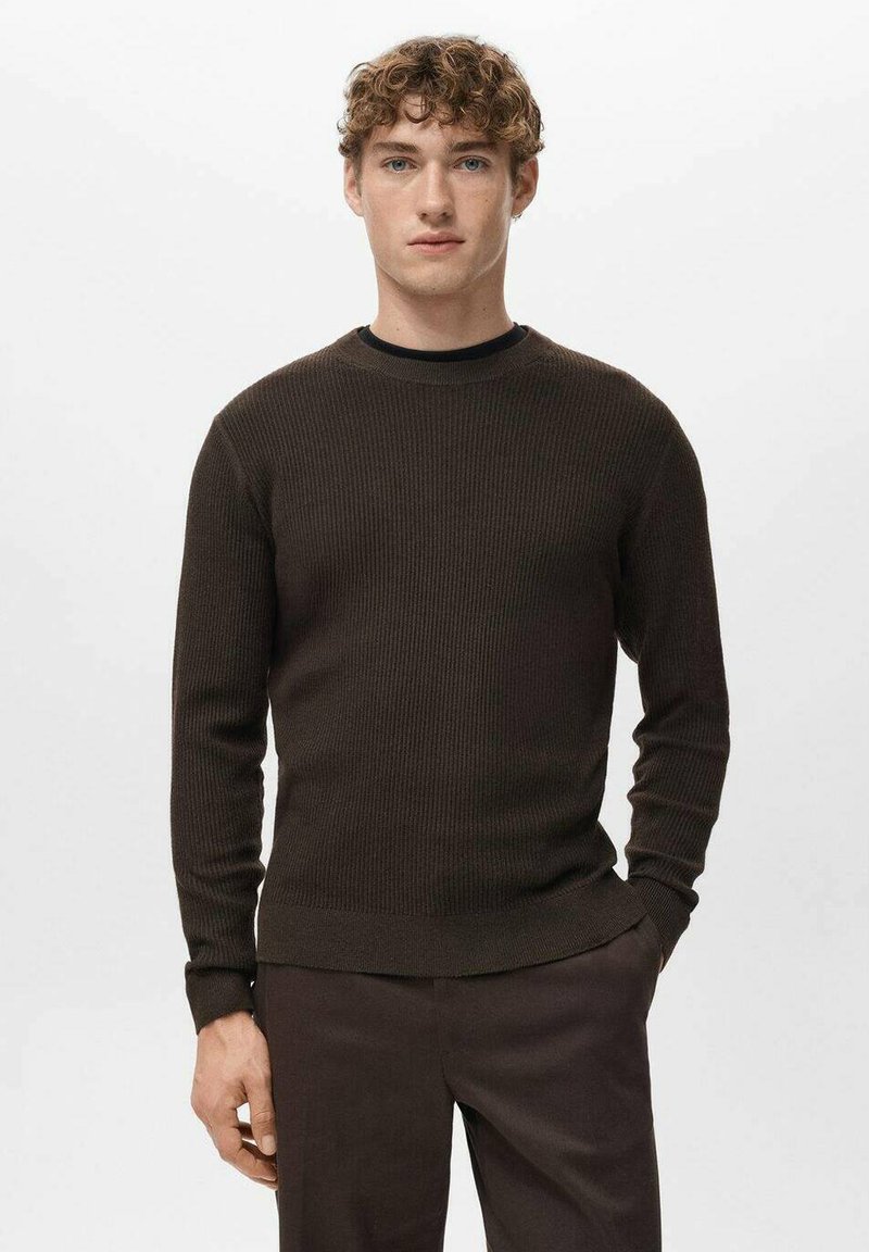 Young man with curly hair wearing a dark brown ribbed sweater and matching pants, standing against a plain light background.