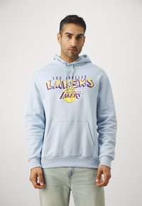 Svijetloplava dukserica s grafikom "Los Angeles Lakers" u ljubičastoj i žutoj boji. Opremljena je prednjim kangaroo džepom i rebrastim manžetama.