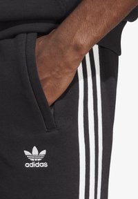 Svarta mjukisbyxor med tre vita ränder längs sidan, utrustade med en sidoficka och en broderad vit Adidas-logotyp på låret.