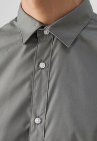 Gros plan d'une personne portant une chemise grise boutonnée avec un col pointu et trois boutons blancs visibles attachés.