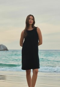 Robe noire sans manches avec un col rond, coupe ample et longueur genou, présentée sur un fond de plage avec des vagues océaniques.