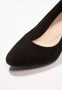 Escarpin en daim noir avec un bout pointu et un design échancré. L'intérieur présente une doublure beige lisse et des détails de couture subtils.