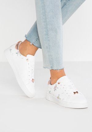 Hvide lave sneakers med lyserødt blomsterprint og sløjfer, båret med lyseblå jeans med frynset kant på hvid baggrund.