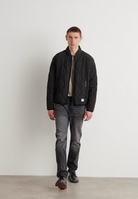 Black quilted bomber jacket with a round neckline, worn over a gray shirt and paired with dark jeans and black shoes. Simple design, no patterns.