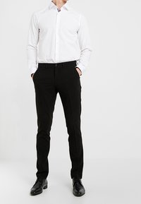 Man som bär svarta byxor, svarta skor och en vit långärmad skjorta med knappar, står med händerna i fickorna framför en neutral bakgrund.