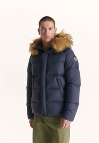 Giubbotto invernale blu navy con cappuccio foderato in pelliccia, dotato di sezioni trapuntate e un patch logo sul lato. Texture liscia, chiusura frontale a zip completa e colletto morbido.