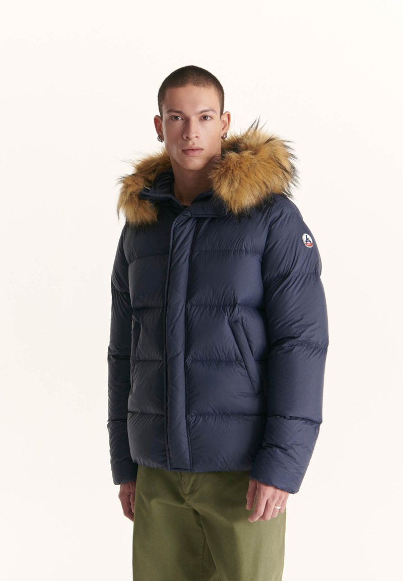 Giubbotto invernale blu navy con cappuccio foderato in pelliccia, dotato di sezioni trapuntate e un patch logo sul lato. Texture liscia, chiusura frontale a zip completa e colletto morbido.