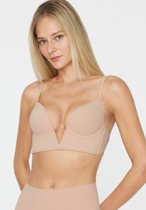 Femme blonde portant un soutien-gorge beige à décolleté plongeant et un bas taille haute assorti, debout devant un fond clair uni.