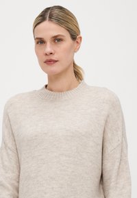 Beige strikkegenser med rund hals, myk tekstur, avslappet passform og senkede skuldre; har subtile variasjoner i garnet for dybde.