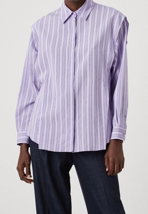 Personne portant une chemise ample à manches longues, rayée verticalement lavande et blanche, et un pantalon foncé, debout devant un fond uni.