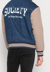 Giubbotto in denim con maniche beige, caratterizzato da un logo bianco "SOCIALITY" e il testo "The Society Since 1990". Polsini e orlo a costine con accenti a strisce.