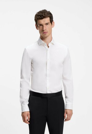 Man met kort, krullend haar, gekleed in een wit overhemd met lange mouwen en een zwarte pantalon, staand tegen een effen lichte achtergrond.