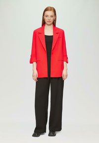 Blazer rouge en tissu lisse, avec un col cranté et un bouton unique, assorti à un pantalon large noir et des chaussures noires ornées.