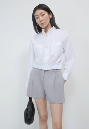 Frau in einem weißen, knopfverzierten, cropped Hemd und grauen, taillierten Shorts, die mit einer Hand eine schwarze Handtasche hält und die andere in der Tasche hat.