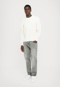 Vit kabelstickad tröja med rund hals, tillsammans med ljusgrå jeans och vita sneakers, stående mot en enfärgad bakgrund.