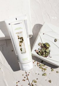 Witte tube van Korres conditioner met groene accenten, voorzien van botanische illustraties en labels. De achtergrond bevat verspreide kruiden op een textuurrijke oppervlakte.
