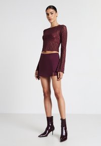 Burgunderfarbenes Spitzen-Longsleeve-Crop-Top mit rundem Ausschnitt, kombiniert mit einem passenden Minirock mit einem Schlitz vorne. Das Model trägt glänzende Stiefeletten.