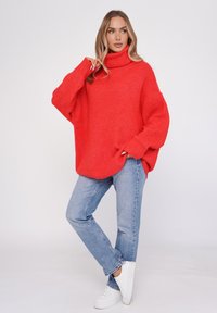 Czerwony, oversize sweter z wysokim kołnierzem, prążkowaną teksturą, długimi rękawami i luźnym krojem, zestawiony z jasnoniebieskimi dżinsami i białymi trampkami.