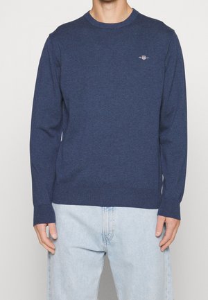 Homme portant un pull à manches longues bleu marine avec un petit logo sur la poitrine et un jean en denim bleu clair.