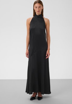 Femme portant une robe longue noire sans manches à col halter avec des chaussures noires à bout pointu, debout sur un sol gris.