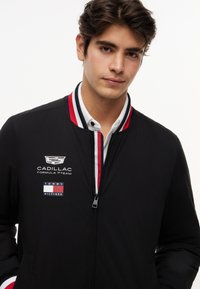 Young man wearing a black Tommy Hilfiger jacket with Cadillac Formula 1 Team logos, white shirt underneath, and red, white, and blue striped collar.