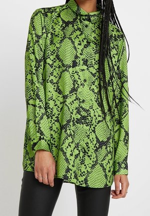 Zielona bluzka z długim rękawem w czarny wzór węża, noszona z czarnymi spodniami, z kołnierzykiem i mankietami zapinanymi na guziki.