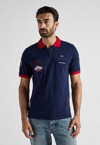 Granatowa koszulka polo z czerwonym kołnierzykiem i wyko ńczeniami rękawów, z naszytymi łatami i zamkiem z przodu. Miękka tkanina i slim fit.