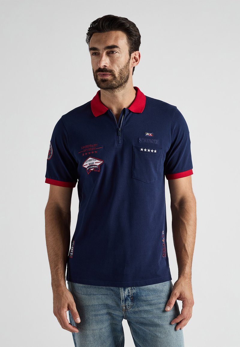 Granatowa koszulka polo z czerwonym kołnierzykiem i wykończeniami rękawów, z naszytymi łatami i zamkiem z przodu. Miękka tkanina i slim fit.