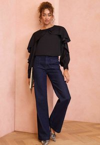 Top nero con maniche a volant, abbinato a pantaloni ampi in denim scuro a vita alta. Tacchi con stampa animale e una clutch dorata completano l'outfit.