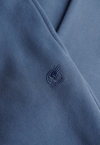 Miękka niebieska tkanina z małym haftowanym ciemnoniebieskim logo skrzydlatej stopy w pobliżu szwu lub zgięcia.