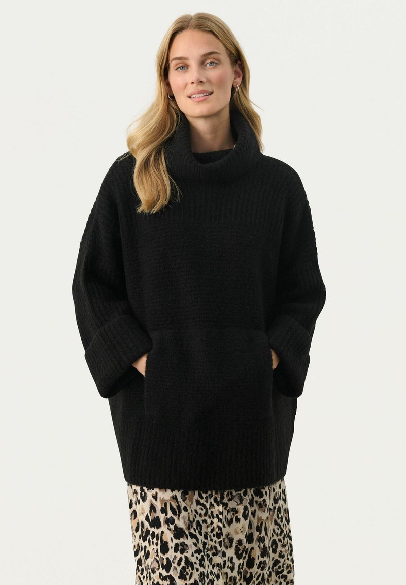 Zwarte oversized coltrui van textuurstof, met geribbelde manchetten en twee opgestikte zakken. Gedragen over een legging met luipaardprint.