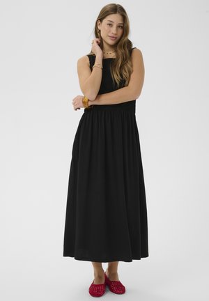 Kvinde iført en sort maxikjole uden ærmer og røde vævede slip-on sko, poserer med armene krydset og langt løst hår.