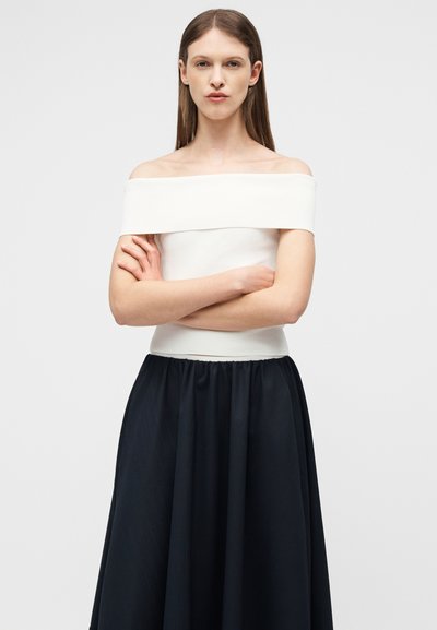 Γυναίκα με μακριά καστανά μαλλιά, με σταυρωμένα τα χέρια, φορώντας ένα λευκό τοπ με ακάλυπτους ώμους και μια μακριά μαύρη φούστα, μπροστά σε ένα απλό φωτεινό φόντο.