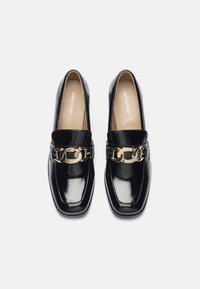 Črne lakirane usnjene loafers s kvadratnim koncem, ki imajo zlati verižni dodatek čez vamp in bež notranjost.