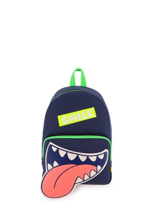 Sac à dos bleu marine avec fermeture éclair verte et poignée, poche avant montrant une bouche ouverte de dessin animé avec des dents et une grande langue rouge, et une étiquette jaune "SMILE".