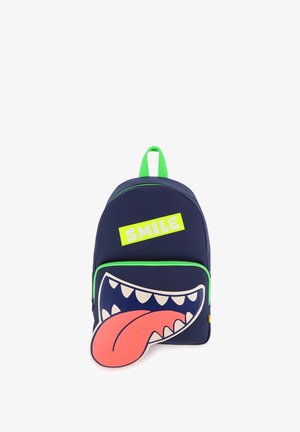 Sac à dos bleu marine avec fermeture éclair verte et poignée, poche avant montrant une bouche ouverte de dessin animé avec des dents et une grande langue rouge, et une étiquette jaune "SMILE".