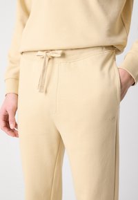 Personne portant un pantalon de survêtement beige avec taille à cordon et poches latérales, une main dans la poche, petit logo "HUGO" sur la cuisse.