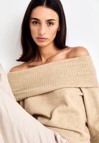 Beige ribbed off-shoulder sweater with wide neckline and soft texture, worn over a light-colored fabric, highlighting the relaxed fit.