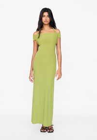 Robe maxi verte avec un décolleté dégageant les épaules, silhouette ajustée, texture douce et fines bretelles. Associée à des sandales noires à lanières.