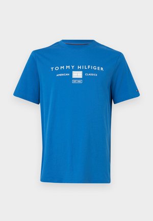 Blå T-shirt med korte ærmer og hvide bogstaver samt logo "Tommy Hilfiger American Classics Est. 1985" trykt på brystet.