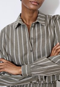 Camisa a rayas verde oliva hecha de un tejido texturizado. Con cuello, cierre de botones y detalle fruncido en la cintura, además de mangas largas.